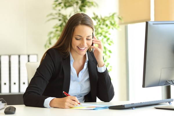 Happy businesswoman calling on mobile phone and taking notes on a desk at office